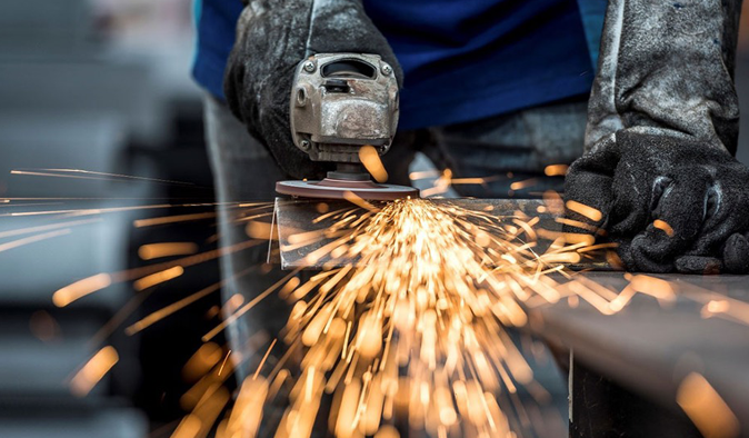 Sparks fly as a glove-clad worker uses an orbital sander to grind a piece of metal in a production setting.
