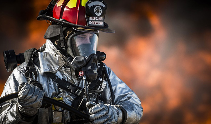 Fire-rescue crew chief holds hose against a blurred fiery backdrop in a protective service.